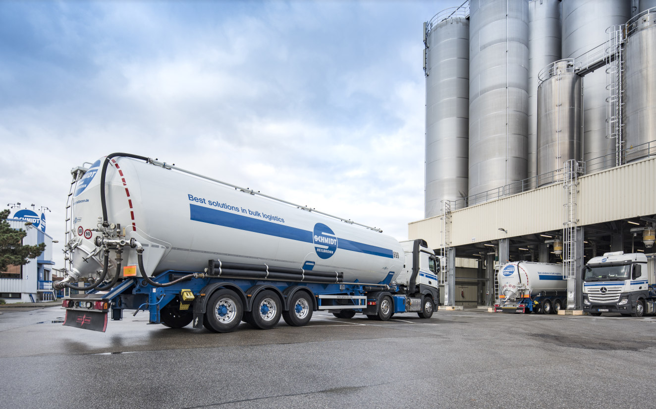 Industrial site with several white tanker trucks positioned in front of large metal storage silos and a production facility. One tanker truck is driving across the paved yard while others are stationed nearby. The scene represents industrial logistics operations supported by stable system operations and continuous application support through SAP AMS.